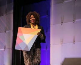 Keynote Mary Spio walks on stage with a cutout of the AnitaB.org logo in her hands during the GHC 17 Thursday Keynote