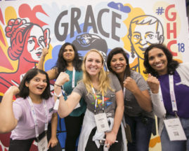 A diverse group of young women stand in front of a mural of Ada Lovelace, Grace Hopper, and Anita Borg