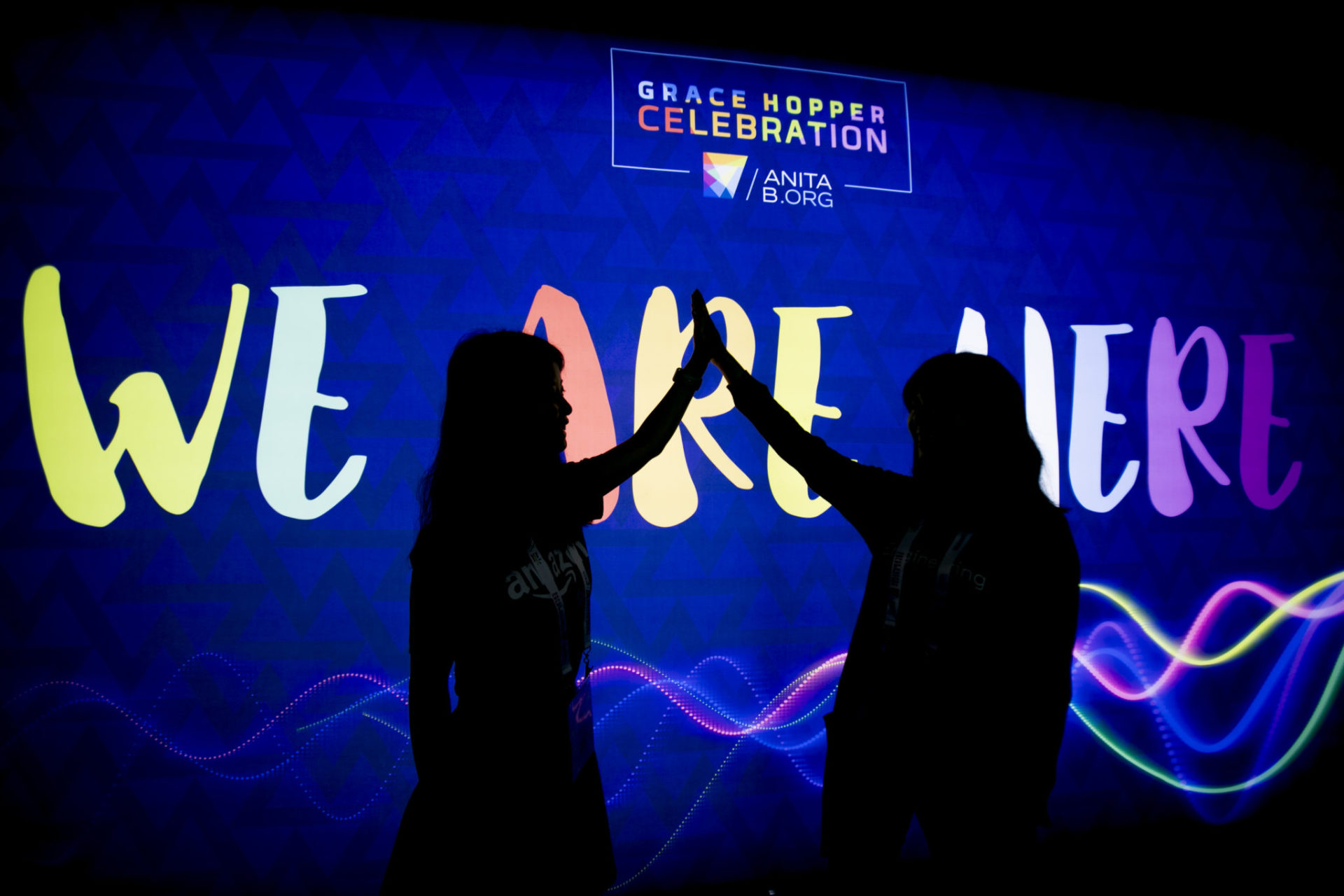 To women highfive in front of a light-up "We Are Here" sign