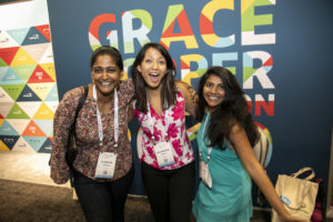Three women stand and smile in front of a Grace Hopper Celebration board at the GHC 19 Expo