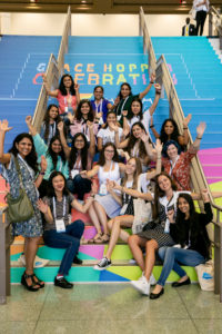 A group of women attendees stand by the stairs with the GHC logo branding at GHC 19