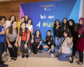 A group of attendees stand in front of a sign that says, "We are at #GHC19"