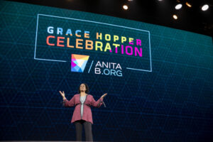 Woman speaking with arms raised with Grace Hopper Celebration sign in the background.